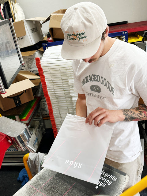 Person wearing a white cap and t-shirt, holding a clear bag with text, in a print shop setting